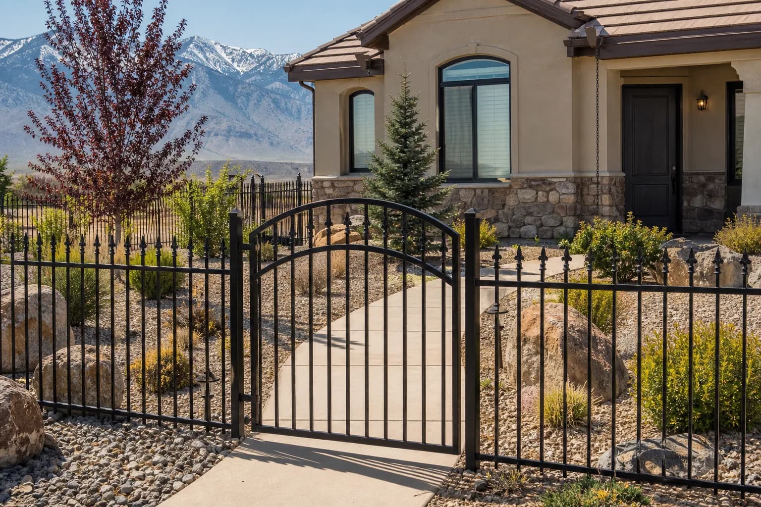 Black ornamental steel picket fence with finials and an arched walk gate in front of a stucco home with desert landscaping